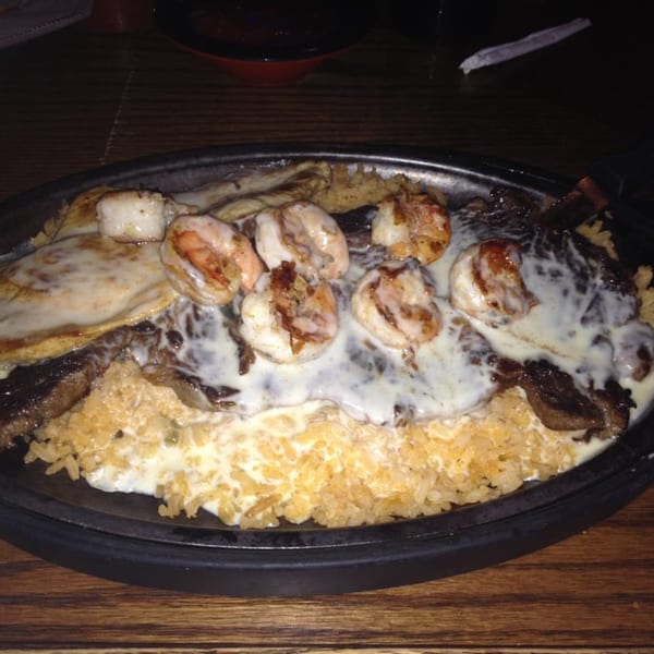Sizzling platter of shrimp, steak, and chicken covered in white cheese sauce and rice.